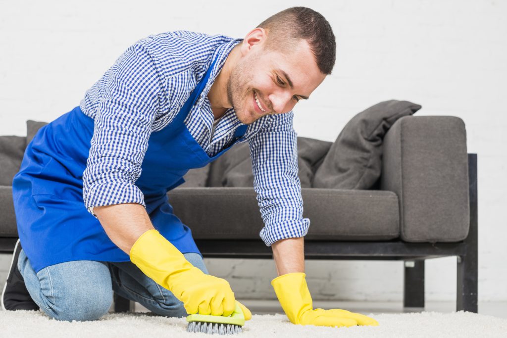 Person-wearing-yellow-protective-gloves-with-a-spray-and-sponge-in-hand-cleaning-a-white-rug